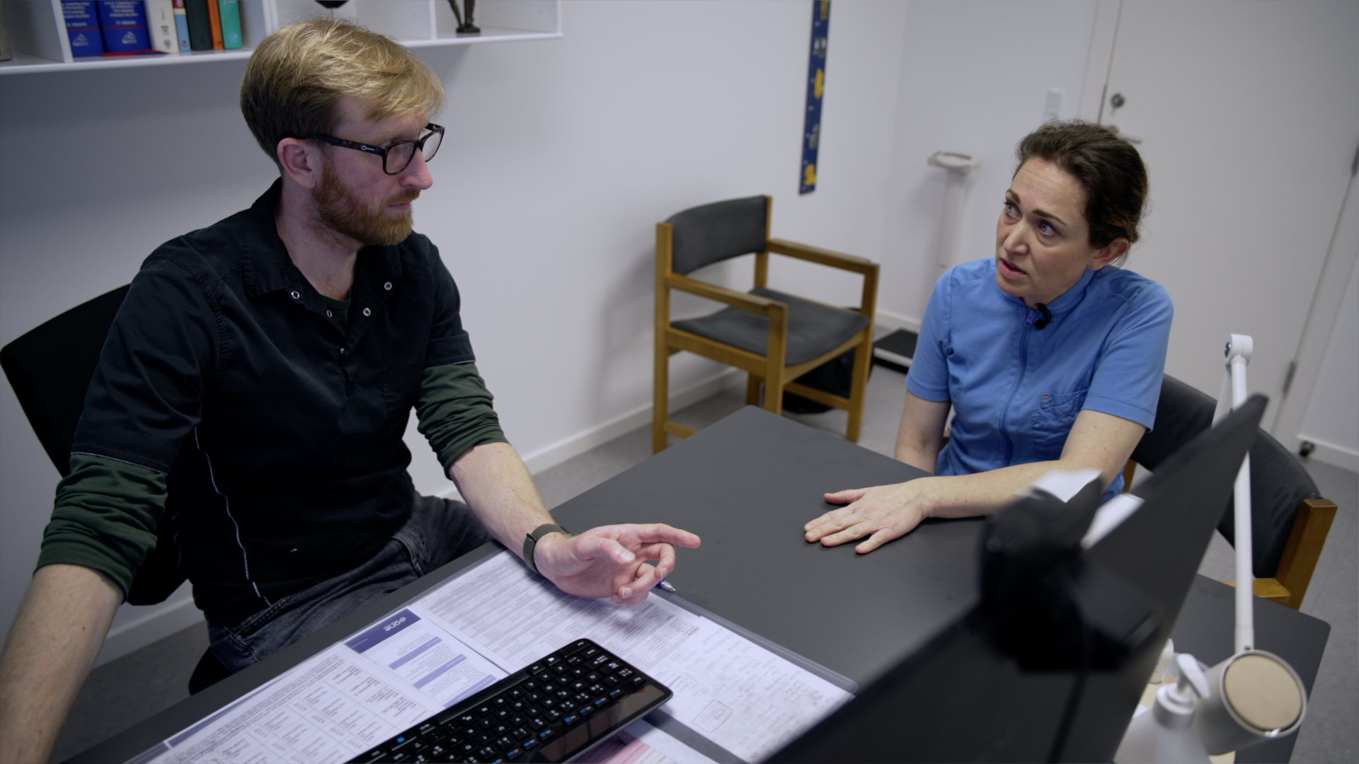 Ung mand og ung kvinde taler sammen ved et skrivebord i en lægepraksis. 