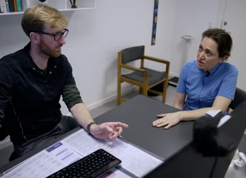 Ung mand og ung kvinde taler sammen ved et skrivebord i en lægepraksis.