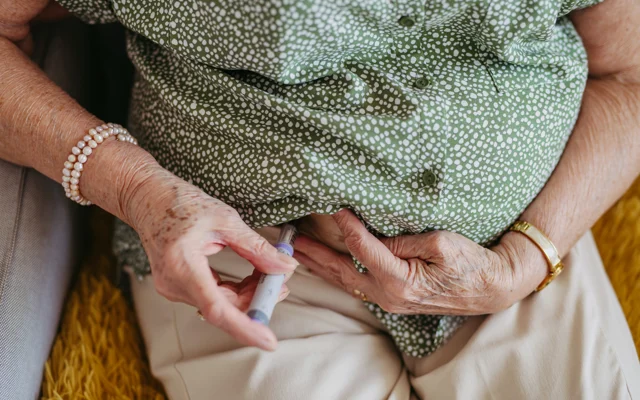 Ældre person sidder og stikker sig i maven med en injektionspen. Ansigt ses og underben ses ikke.