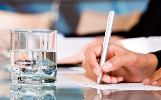 Hånd skriver på papir med et glas vand i forgrunden