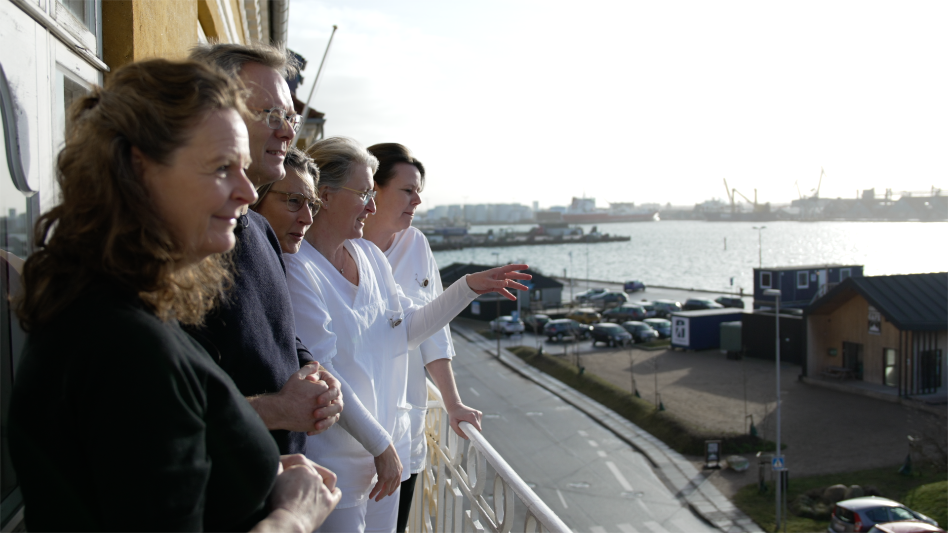 Fem personer står smilende på en altan i solskin og kigger ud over, hvad man må gætte er en havn og en by. Det er solskin.