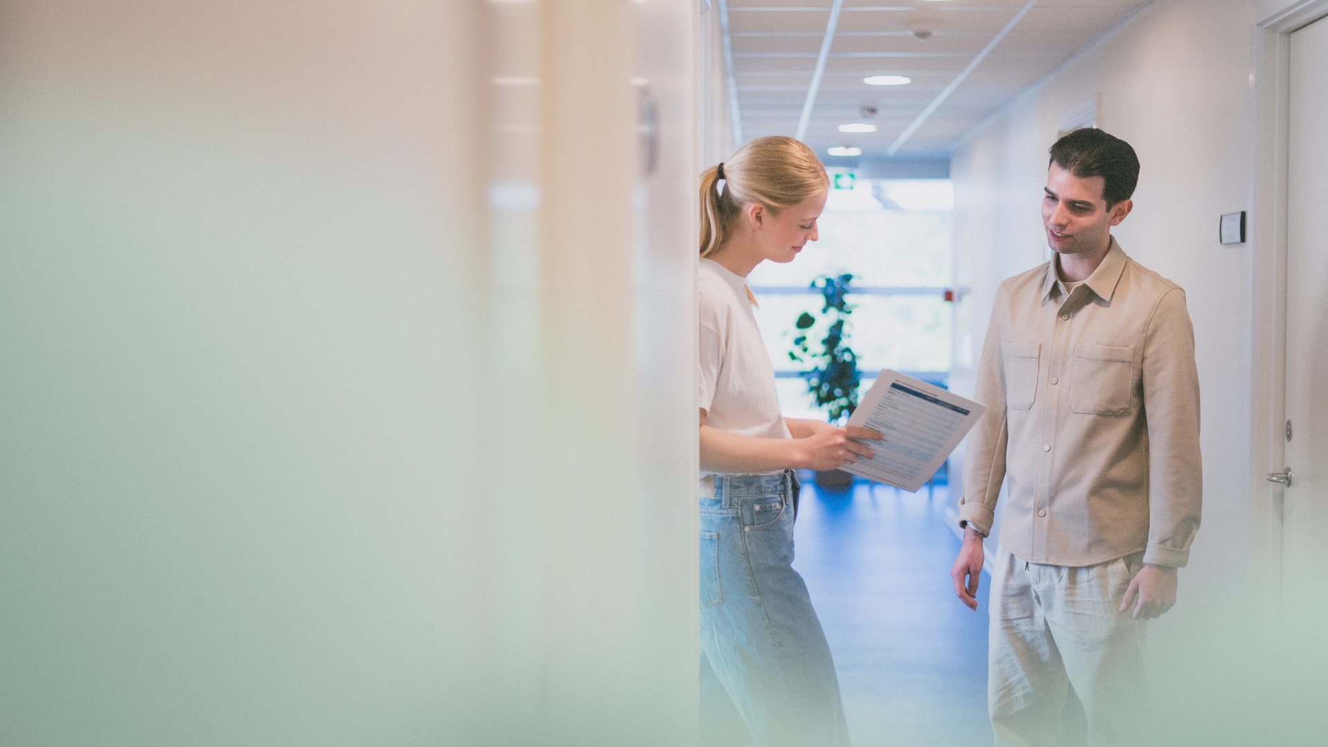 To unge står i en gang i en lægeklinik og taler med hinanden: Den ene peger på et stykke papir