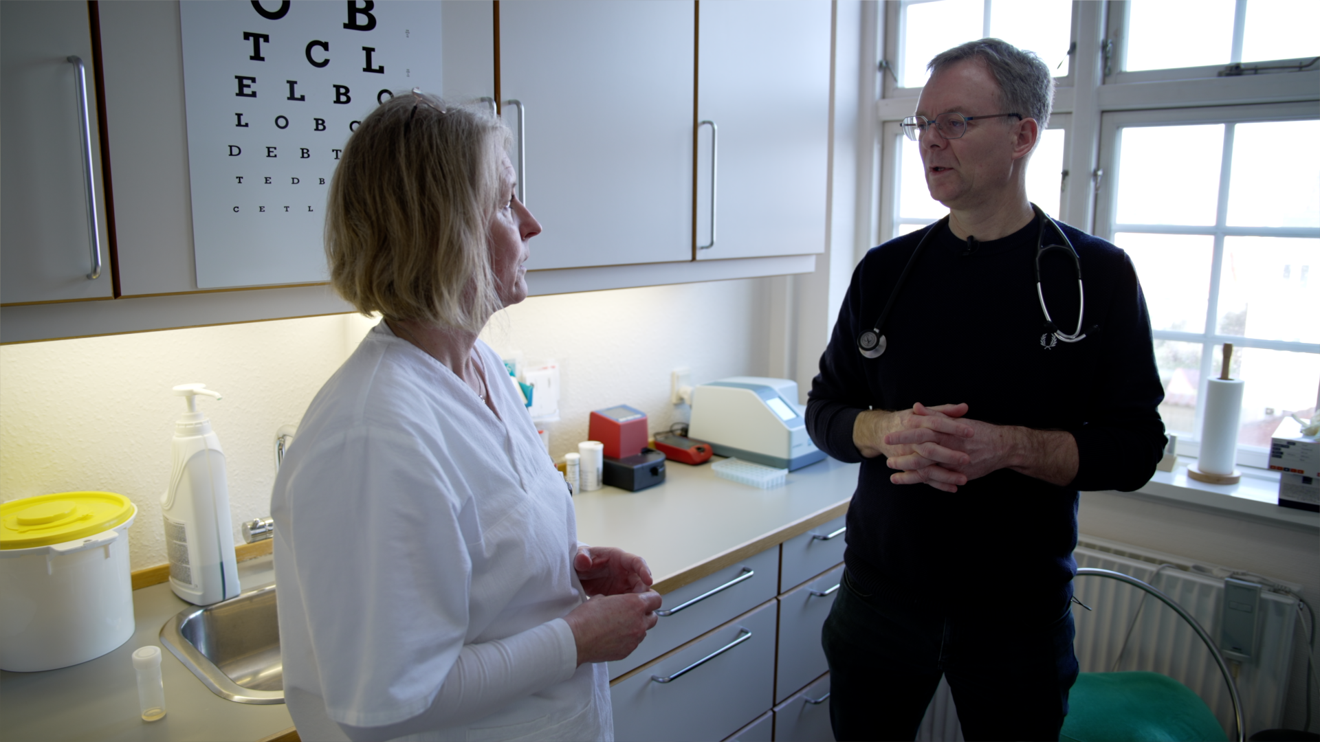 Kvinde og mand taler sammen i et køkkenlignende rum med laboratorieudstyr foran et vindue.