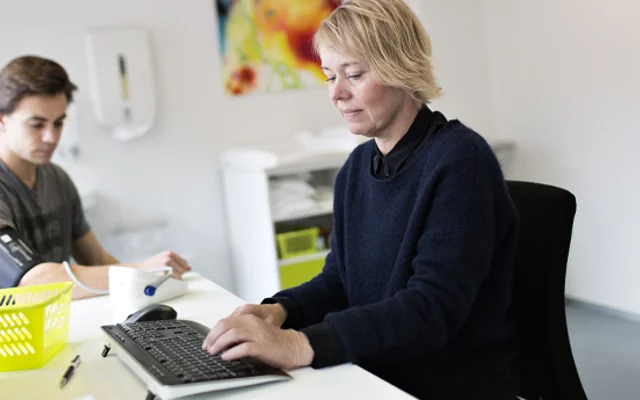 Læge ved computeren. Patient ved siden af, der får taget blodtryk