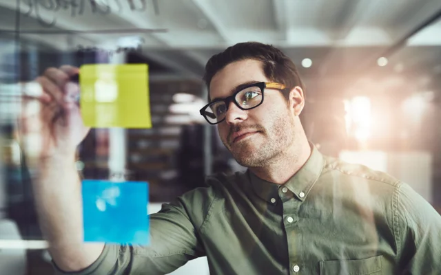 Person markerer noget på en post-it, som er hængt på glasvæg ved siden af en anden i et kontorlokale