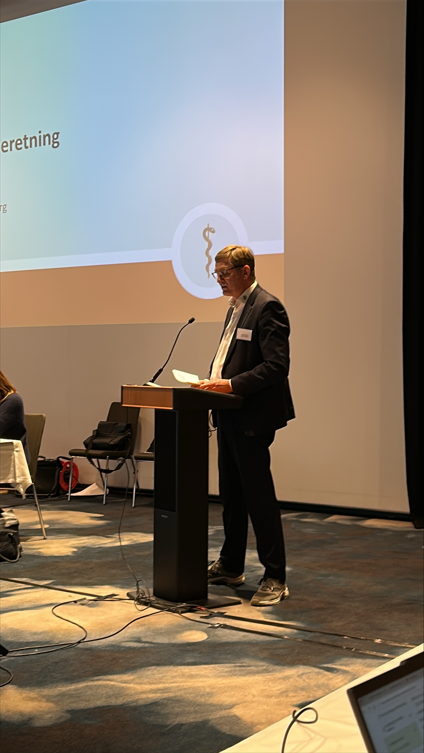 Mand holder tale ud fra manus foran storskærm