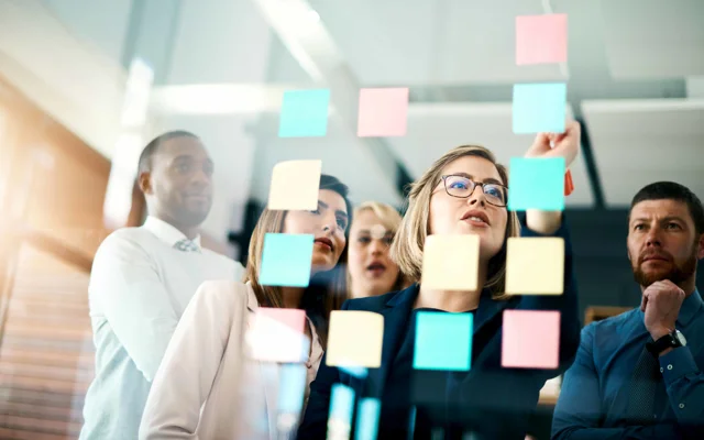 En gruppe mennesker ses ved glasvæg kiggende på post-its som en af dem skriver på.