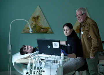 Person ligger på briks i en lægekonsultation. Ung læge kigger på skærm. Bag hende står ældre mandlig læge