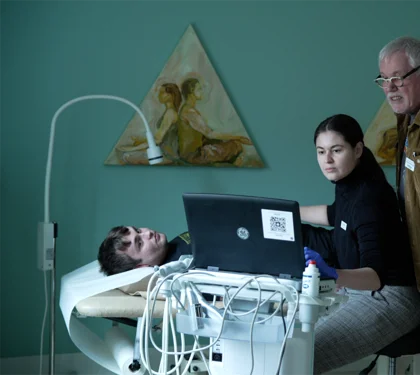 Person ligger på briks i en lægekonsultation. Ung læge kigger på skærm. Bag hende står ældre mandlig læge 